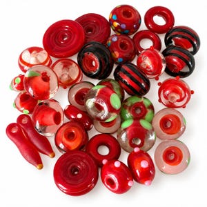 May include: A collection of assorted glass beads in various shades of red, featuring clear, opaque, and translucent finishes. The beads include round, donut, and cylindrical shapes, some decorated with black stripes, green dots, or multicolored spots.