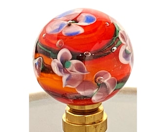 May include: A decorative lamp finial with a vibrant, hand-blown glass orb. The orb features a fiery red and orange base with delicate white and purple floral accents. The finial is mounted on a brass base, adding a touch of elegance.