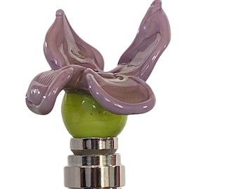 May include: A decorative bottle stopper featuring a lavender glass flower design atop a green glass sphere. The flower has detailed petals and sits on a silver metal base. A unique accent for any bar.
