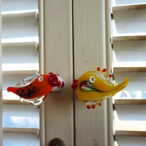 May include: Two colorful glass fish door knobs. One is orange with black spots and a white and red bulbous end. The other is yellow with red accents. Both have a whimsical design and are mounted on a white door.