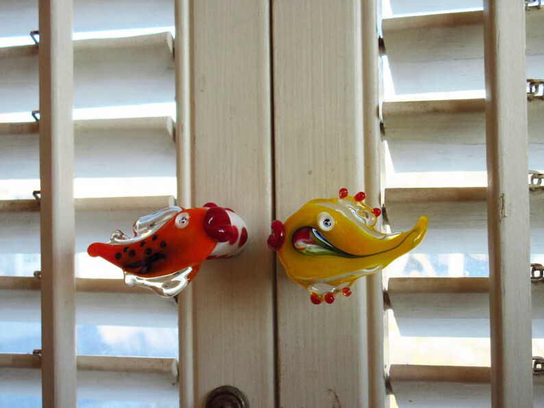 May include: Two colorful glass fish door knobs. One is orange with black spots and a white and red bulbous end. The other is yellow with red accents. Both have a whimsical design and are mounted on a white door.
