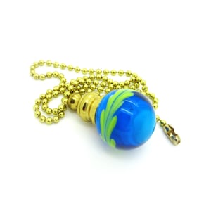 Könnte beinhalten: Ein Deckenventilator-Zugkette aus blauem und grünem Glas mit einem messingfarbenen Metallsockel und einer Kette. Die Glaskugel hat ein Wirbeldesign mit Blau- und Grüntönen. Die Kette besteht aus kleinen, runden Perlen.
