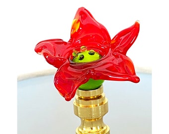 May include: A red glass flower lamp finial with a green center and gold base. The flower has a textured, ruffled appearance.