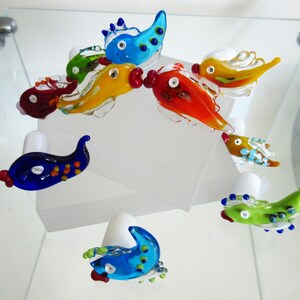 May include: A collection of colorful, handcrafted glass fish figurines. The fish are in various colors, including blue, green, yellow, orange, and red, with white accents. Each fish has a unique design and is displayed on a white surface.
