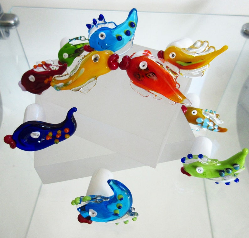 May include: A collection of colorful, handcrafted glass fish figurines. The fish are in various colors, including blue, green, yellow, orange, and red, with white accents. Each fish has a unique design and is displayed on a white surface.