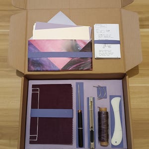 Könnte beinhalten: Eine offene Pappschachtel mit Buchbinderei-Zubehör. Darin befinden sich ein Stapel Papiere, ein Heft, ein burgunderfarbenes, mit Stoff bezogenes Buch, eine Nadel, ein Cutter, Faden, ein Falzbein und andere Werkzeuge. Die Schachtel ist mit lila Papier ausgekleidet.