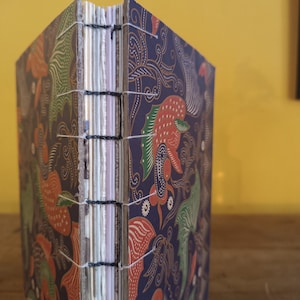 May include: A handcrafted journal with a batik-style cover. The cover displays a dark blue background with vibrant floral and bird patterns in red, green, and gold. The pages are bound with white thread and black stitching.
