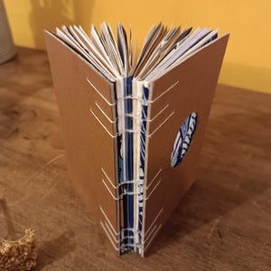 Op de afbeelding: Handgemaakt notitieboek met een bruine kaft en witte stiksels. De pagina's zijn zichtbaar en de randen zijn ruw afgesneden. Een blauw-wit patroonstof is zichtbaar door een cirkelvormige uitsparing op de kaft. Het notitieboek is open.