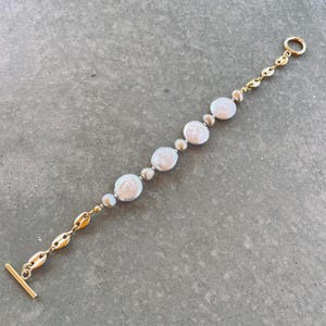 May include: A gold-toned bracelet featuring large, flat, off-white pearls interspersed with smaller, round, gold-colored beads. The bracelet has a toggle clasp and a chain-link design on each end. The pearls have a slightly iridescent sheen.