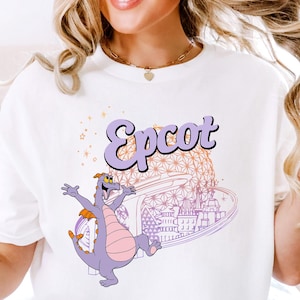 May include: White t-shirt featuring the word "Epcot" in a purple, retro font. A cartoon dragon in purple and pink is depicted with arms outstretched, in front of a stylized image of a theme park. The shirt is a comfortable, casual style.