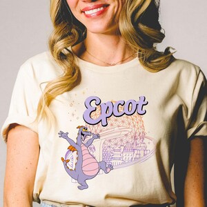 May include: Cream-colored t-shirt featuring the word "Epcot" in a purple, retro font. The shirt displays a cartoon dragon character and a stylized image of a theme park. The design is in shades of purple, pink, and orange.