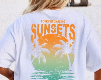 May include: White t-shirt with a sunset graphic. The design features a vibrant orange and green sunset scene with palm trees and the words "FOREVER CHASING SUNSETS". The shirt is worn on a beach.