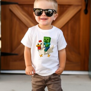May include: White t-shirt featuring a pixelated graphic of a green zombie riding a white chicken. The chicken has a red comb and yellow feet. The child is wearing sunglasses and brown pants.