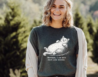 May include: Dark green t-shirt featuring a detailed illustration of a relaxed, smiling cat. The cat is lying on its back. The text "MOTHER, I AM BUT SKIN AND BONES." is printed below the cat. The person is wearing a light-colored cardigan.