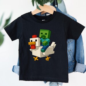 May include: Black t-shirt featuring a pixelated graphic of a green zombie riding a white chicken. The chicken has a red comb and yellow feet. The design is in a cartoon style, perfect for kids.