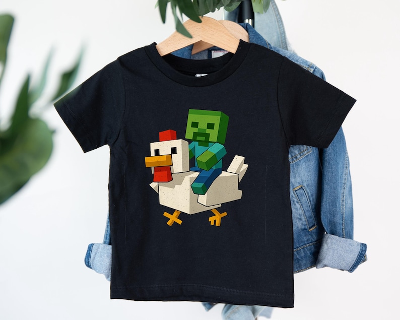May include: Black t-shirt featuring a pixelated graphic of a green zombie riding a white chicken. The chicken has a red comb and yellow feet. The design is in a cartoon style, perfect for kids.