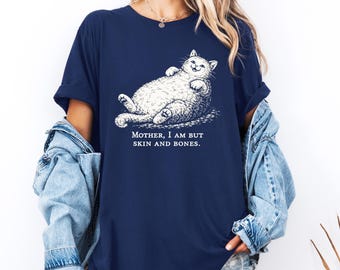 May include: Navy blue t-shirt featuring a whimsical illustration of a relaxed cat. The cat is lying on its back with its paws in the air. The text "MOTHER, I AM BUT SKIN AND BONES" is below the cat.