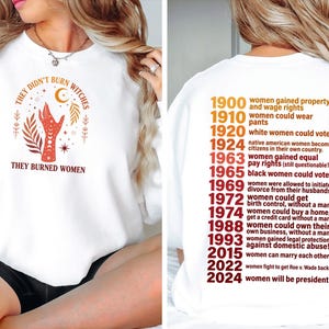 May include: A white crewneck sweatshirt featuring a front graphic of a hand with celestial symbols and the text 'THEY DIDN'T BURN WITCHES THEY BURNED WOMEN'. The back displays a chronological list of historical milestones in red and orange text.