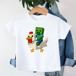 May include: White short-sleeved t-shirt featuring a pixelated green zombie riding a pixelated white chicken. The zombie has blue arms and legs. The chicken has a red comb and yellow feet. The design is centered on the shirt.