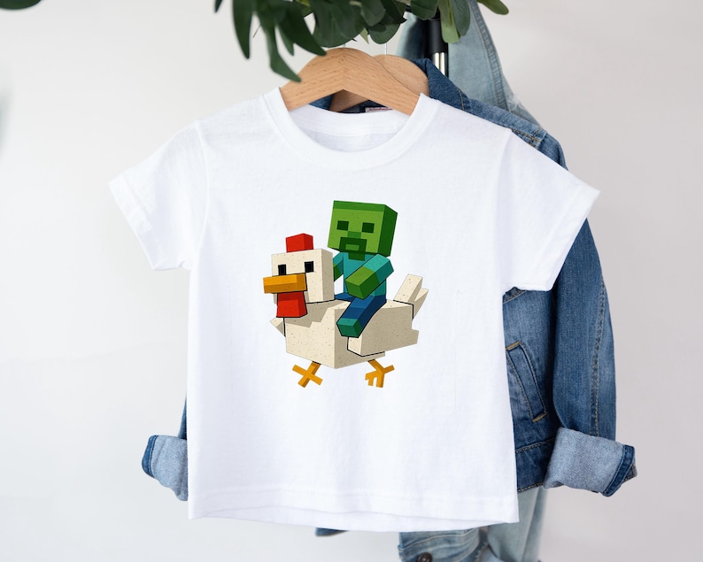 May include: White short-sleeved t-shirt featuring a pixelated green zombie riding a pixelated white chicken. The zombie has blue arms and legs. The chicken has a red comb and yellow feet. The design is centered on the shirt.