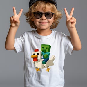 May include: White t-shirt featuring a pixelated design of a zombie riding a chicken. The zombie is green and blue, and the chicken is white with red and yellow accents. The person is wearing sunglasses and a hat, and is giving the peace sign.