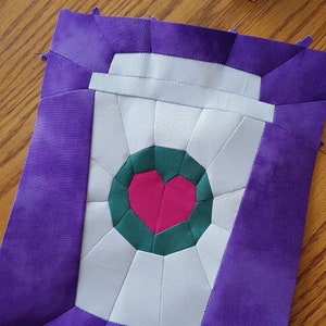 May include: A quilted coffee cup design with a heart in the center. The cup is white with a green and pink heart. The background is purple. The design is made of fabric and stitched together.