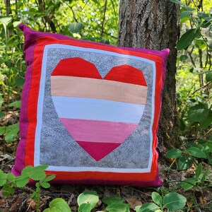 May include: A pink, white, red, and gray heart-shaped pillow with a gray background. The pillow is surrounded by green foliage.