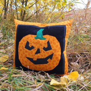 Op de afbeelding: Een handgemaakt oranje en zwart kussen met een jack-o'-lantern ontwerp. Het kussen ligt op een bed van droog gras en herfstbladeren.