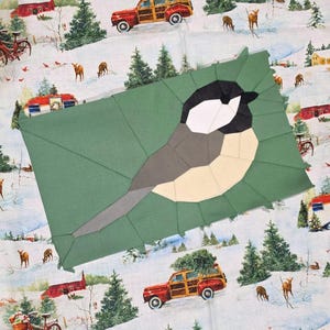 May include: A rectangular fabric quilt block featuring a pieced chickadee design in black, white, and gray against a solid sage green background, layered over a winter-themed fabric printed with red station wagons and deer.