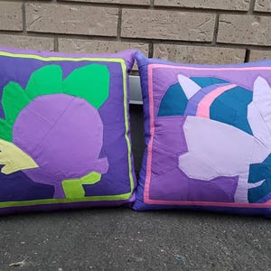 May include: Two purple and green pillow covers with a cartoon design. One pillow features a purple and green dragon, the other features a purple and pink unicorn.