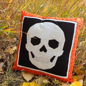 May include: A white skull cushion with a black background and an orange border. The cushion is lying on a bed of brown and green grass with yellow leaves.