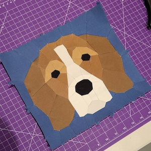 Può includere: Primo piano di una testa di cane in tessuto patchwork marrone e bianco, su uno sfondo in tessuto blu. Gli occhi del cane sono neri e il naso è nero. Il tessuto è cucito insieme in un motivo geometrico.