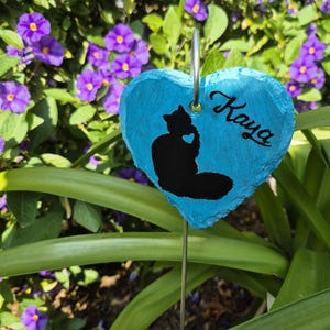 Op de afbeelding: Een blauwe, hartvormige tuinmarker met een zwart katten silhouet en de naam "Kaya" in cursief. De stenen marker heeft een metalen lus. De achtergrond toont groene bladeren en paarse bloemen.