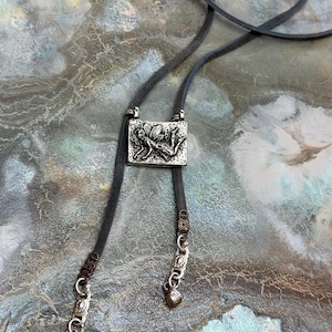 Op de afbeelding: Een zilveren ketting met een gedetailleerd fee-ontwerp hangt aan een bruin leren koord met zilveren kralen en bedels. De ketting wordt getoond op een getextureerd, grijs en bruin oppervlak.