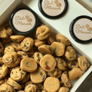 Peut inclure: Une boîte blanche remplie de nombreuses petites pépites de chocolat rondes. Trois contenants noirs circulaires sont placés en haut, chacun avec une étiquette beige portant le texte Dolci Moments et un petit graphique d'un biscuit et d'une part de gâteau.