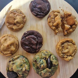 Op de afbeelding: Een bovenaanzicht van een houten snijplank met een assortiment versgebakken koekjes. De koekjes variëren in kleur en textuur, waaronder chocolade en karamel, met zichtbare vullingen zoals koekjes en room.
