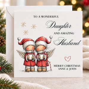 May include: A square white greeting card featuring two illustrated figures with angel wings, wearing red coats and Santa hats, holding mugs with candy canes. The text reads: TO A WONDERFUL Daughter AND AMAZING Husband MERRY CHRISTMAS ANNE & JOHN.
