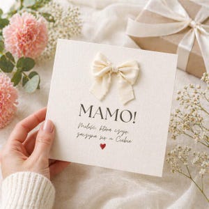 May include: A cream-colored greeting card with a cream-colored bow attached. The card has the word "MAMO!" printed on it, along with additional text and a small red heart. The card is held by a person's hand, with pink flowers and a gift box in the background.