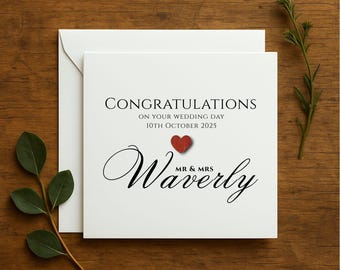 May include: A white wedding card with the words "Congratulations" and "Mr & Mrs Waverly" in black script. A small red heart is above the name. The card is on a wooden surface with greenery.