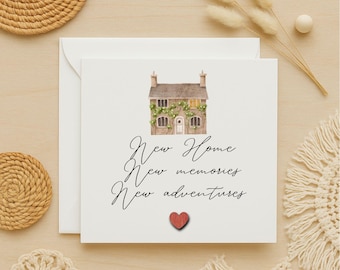 May include: A white greeting card with a watercolor illustration of a house covered in greenery. The card reads "New Home, New memories, New adventures" with a small red heart. The card is on a light wooden surface.