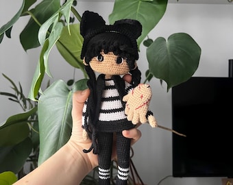 May include: Crocheted doll with black hair, a black bow, and a black and white striped shirt. The doll is holding a small, tan, stitched hand. The doll is wearing black pants and black shoes with white stripes. The doll is a handmade item.