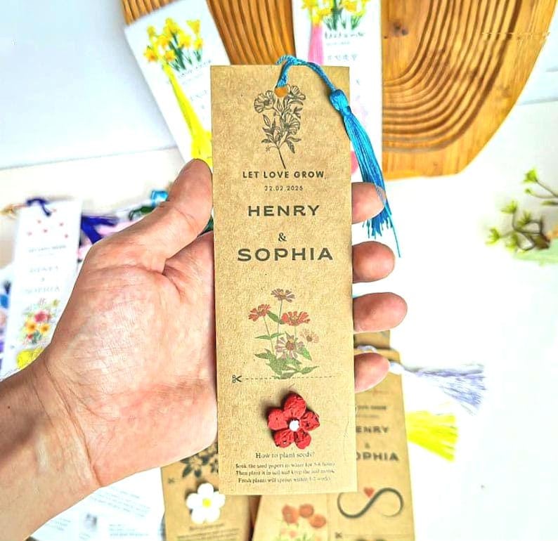 A brown paper bookmark with a floral design and the words 'LET LOVE GROW' and 'HENRY & SOPHIA' printed on it. A red flower is attached to the bookmark. A blue tassel hangs from the top. The bookmark is held by a hand.