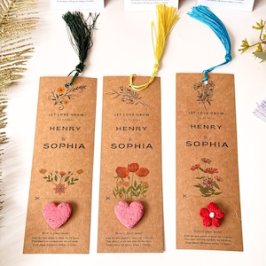 Three brown paper bookmarks with colorful tassels. Each bookmark features the words 'LET LOVE GROW' and the names 'HENRY & SOPHIA' with a date. One has a pink heart-shaped seed, another has red flowers, and the third has a floral design.