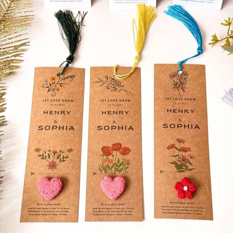 Three brown paper bookmarks with colorful tassels. Each bookmark features the words 'LET LOVE GROW' and the names 'HENRY & SOPHIA' with a date. One has a pink heart-shaped seed, another has red flowers, and the third has a floral design.