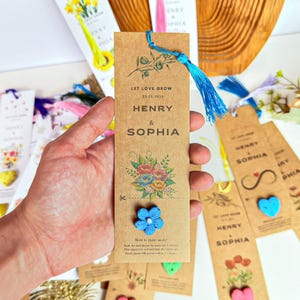 A collection of personalized wedding favors. The favors are brown paper bookmarks with a blue tassel. The bookmarks have the names 'Henry & Sophia' and the date '22.02.2025' printed on them. The bookmarks are decorated with floral designs and a blue flower.