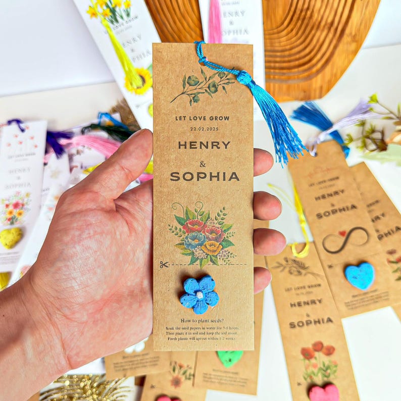 A collection of personalized wedding favors. The favors are brown paper bookmarks with a blue tassel. The bookmarks have the names 'Henry & Sophia' and the date '22.02.2025' printed on them. The bookmarks are decorated with floral designs and a blue flower.