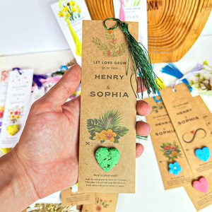 A brown bookmark with the words 'LET LOVE GROW' and the date '22.02.2025' printed on it. The bookmark also has the names 'HENRY & SOPHIA' printed on it. The bookmark has a green heart-shaped seed paper attached to it. The bookmark has a green tassel attached to the top.