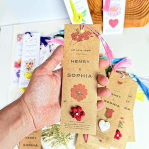 A collection of brown paper bookmarks with pink tassels. The bookmarks feature the names 'Henry & Sophia' and the phrase 'Let Love Grow' along with a date. Each bookmark has a red flower design and instructions for planting seeds. Additional bookmarks with hearts and flowers are visible.