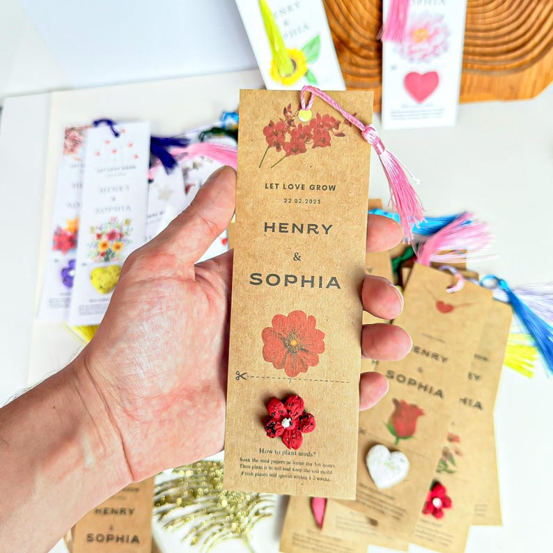 A collection of brown paper bookmarks with pink tassels. The bookmarks feature the names 'Henry & Sophia' and the phrase 'Let Love Grow' along with a date. Each bookmark has a red flower design and instructions for planting seeds. Additional bookmarks with hearts and flowers are visible.