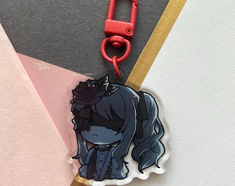 Op de afbeelding: Een zwart-witte anime-achtige sleutelhanger met een rood clipje. Het personage heeft lang, donkerblauw haar, een zwart blinddoek en een zwarte roos op zijn hoofd. Het personage draagt een witte jurk met een zwart lint.